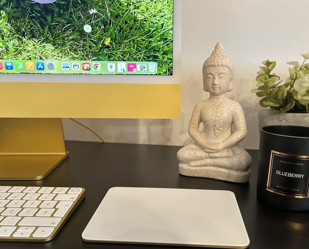 žuti imac na crnom stolu sa tipkovnicom i trackpadom, pored je teglica sa zelenim umjetnim cvijećem, svijećom u crnoj čaši s natpisom i kip bude, slika je simbolična za blog članak u kojem je glavna tema seo optimizacija